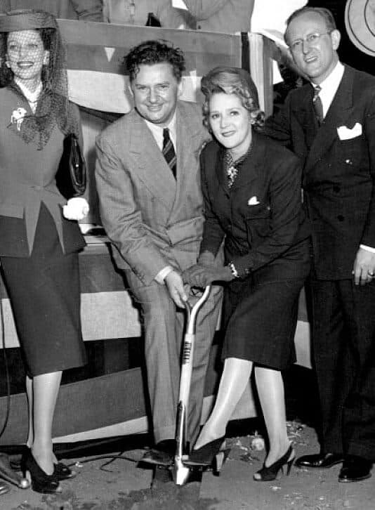 Four people in formal attire are gathered for a groundbreaking ceremony; a woman in the center holds a shovel.