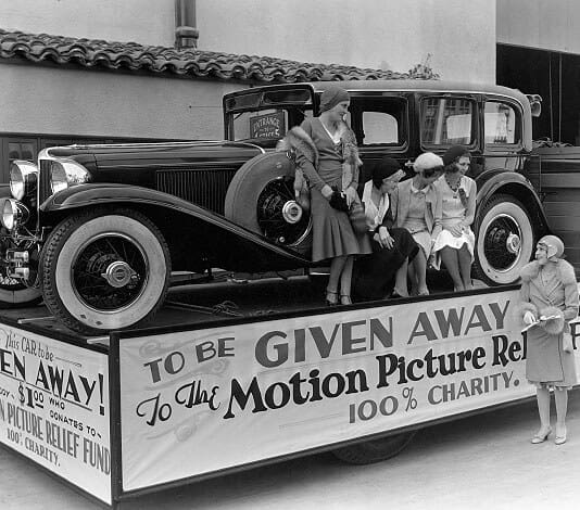 A vintage car on a decorated platform with four women sitting on the car and one standing, promoting a charity giveaway for the Motion Picture Relief Fund.