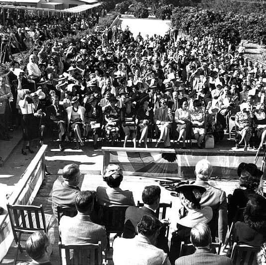 A large crowd seated outdoors, viewed from behind a group of speakers. The event appears formal, with people dressed in suits and dresses, and a photographer capturing the moment.