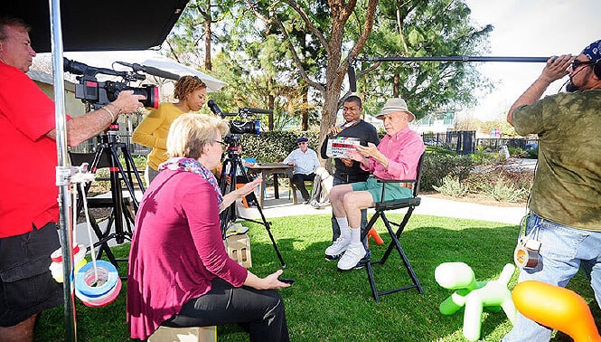 A group of people are filming an outdoor interview. Two men are seated in director's chairs, one holding a tablet, while a camera crew works around them. Bright, sunny day with trees in the background.