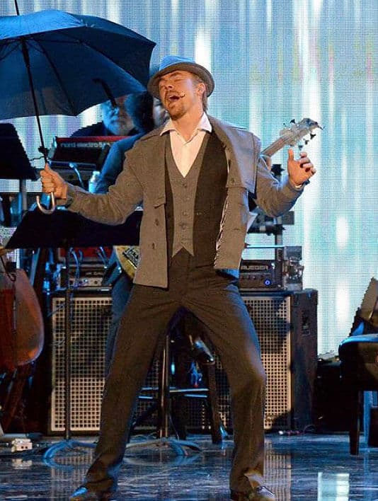 A man is performing on stage, singing into a microphone while holding a black umbrella. He wears a suit and a hat. Musicians with instruments are visible in the background.