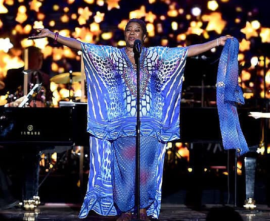 A singer stands on stage with arms outstretched, wearing a blue patterned dress and headband, performing in front of a backdrop with star-shaped lights. A piano and musicians are visible in the background.