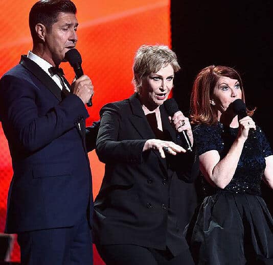 Three people stand on stage, each holding a microphone. The backdrop is red, and they appear to be speaking or performing. The person in the middle gestures with their hands.