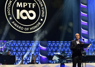 Man speaking on stage at an event with a banner reading "MPTF 100 Award of Honor" in the background and musicians in the setup.