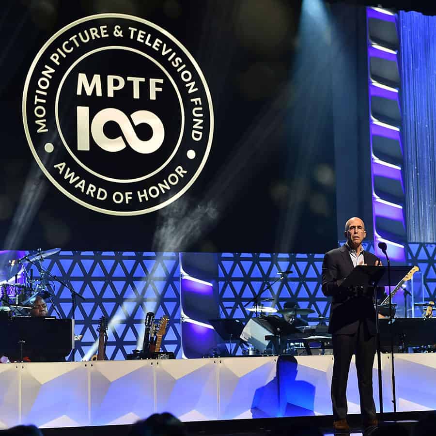 Man speaking on stage at an event with a banner reading "MPTF 100 Award of Honor" in the background and musicians in the setup.