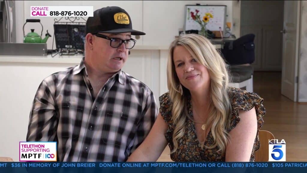 A man wearing a black and yellow cap sits next to a woman with long blonde hair; both appear on a telethon broadcast with informational text and donation numbers displayed on the screen.