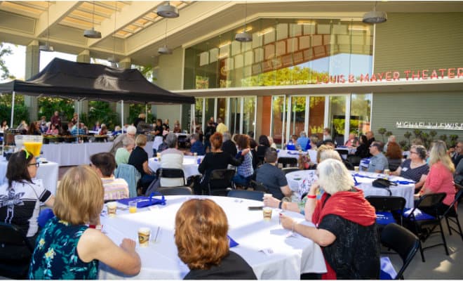 Outdoor community event with people seated at tables, listening to a speaker under a canopy.