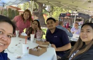 Five people sitting at a table under an outdoor canopy, smiling at the camera. There are food containers and drinks on the table. People and a food truck are visible in the background.