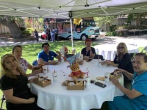 A group of six people sits around a table under a canopy, enjoying a meal with food boxes and drinks, with a blue and black food truck visible in the background. A teddy bear is on the table.