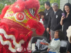 A child smiles and holds a red envelope while interacting with a performer in a vibrant red and yellow lion costume. Several adults observe and take photos in the background.