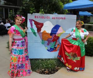 Two women in traditional Mexican dresses stand beside a colorful map of Mexico labeled "Republica Mexicana" outdoors. Both are wearing green sashes and smiling at the camera.