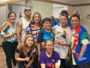 A group of eight people posing for a photo indoors, some holding pride flags and wearing colorful clothing and accessories, smiling towards the camera.