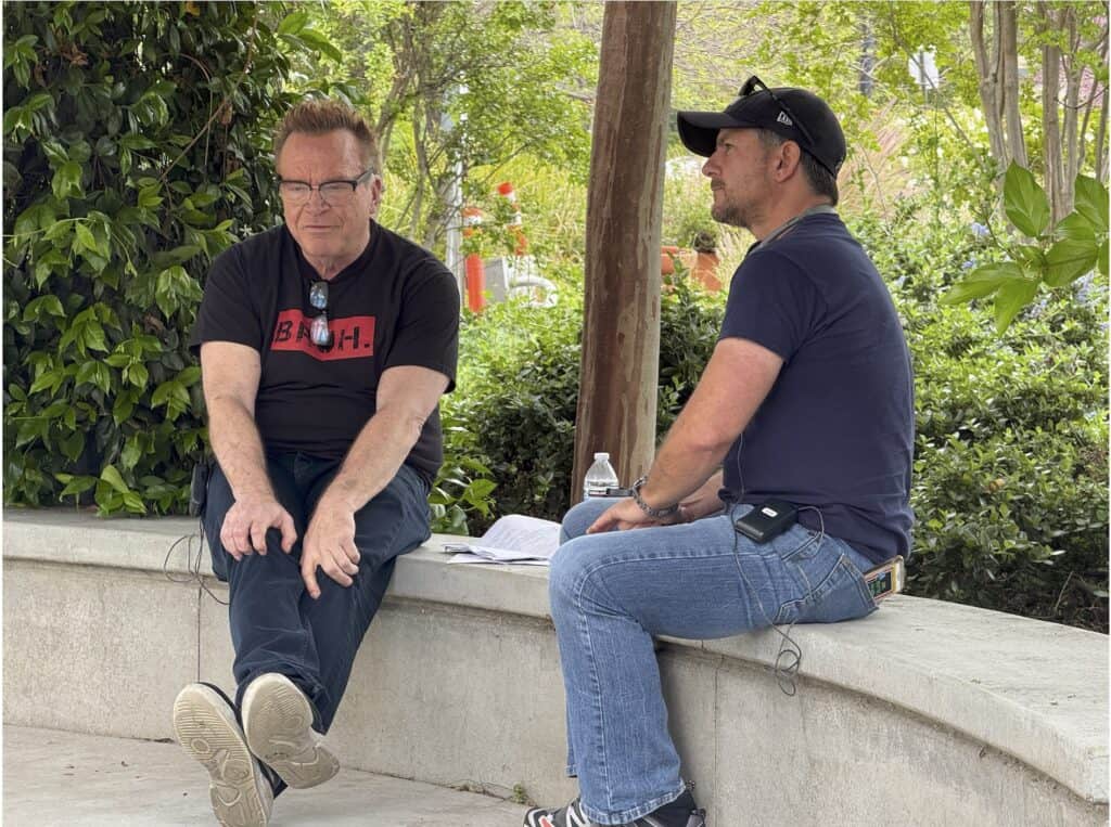Two men sit and talk on a curved concrete bench outdoors, surrounded by greenery. One man holds papers, and a water bottle is placed beside them.