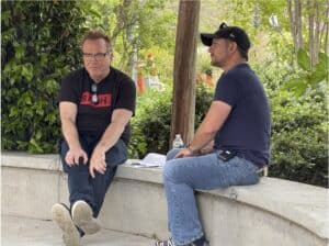 Two men sit and talk on a curved concrete bench outdoors, surrounded by greenery. One man holds papers, and a water bottle is placed beside them.