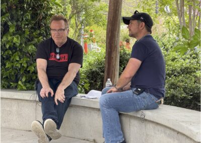 Two men sit and talk on a curved concrete bench outdoors, surrounded by greenery. One man holds papers, and a water bottle is placed beside them.